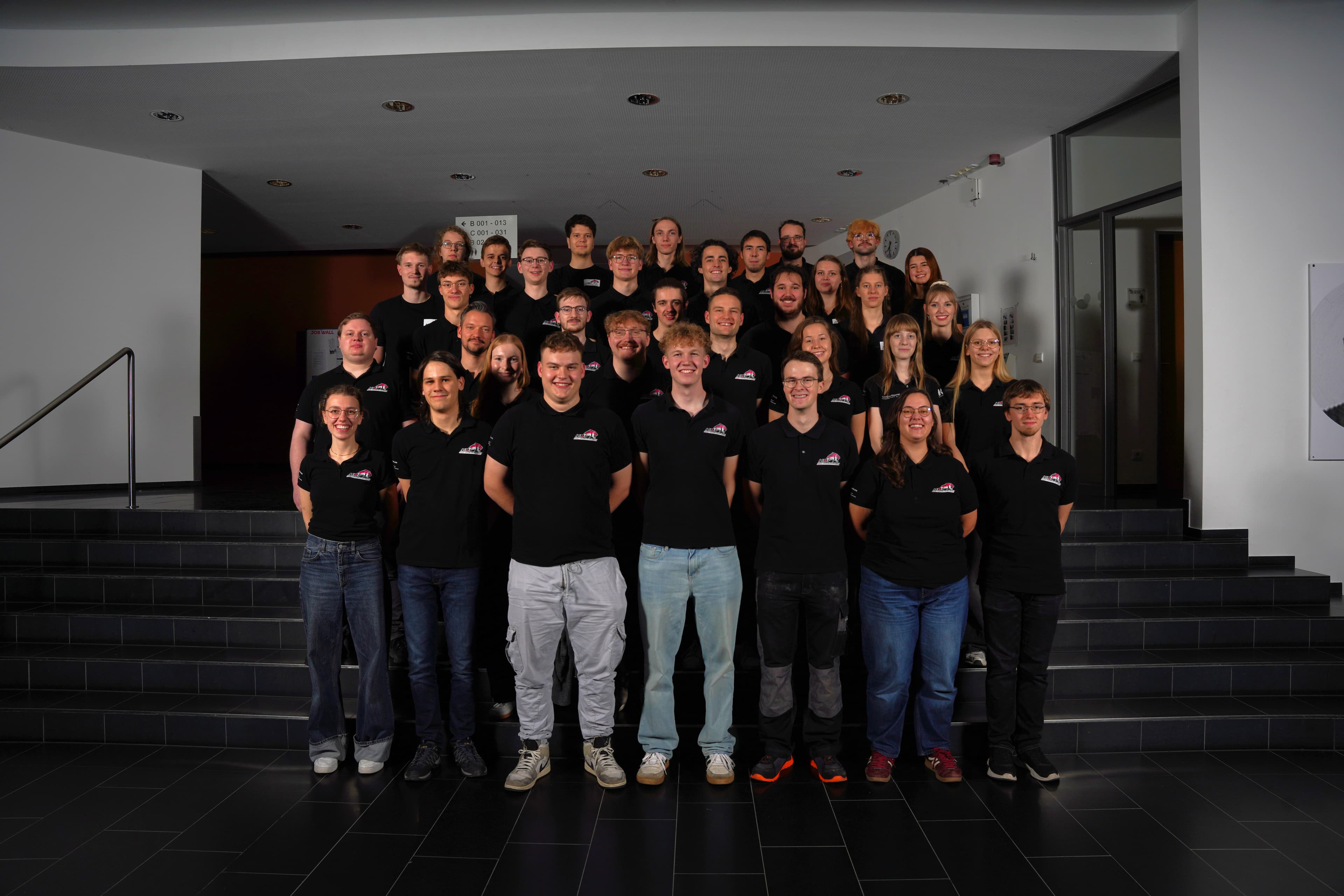 Teammitglieder 2025/26, lächelnd auf Treppenstufen der Hochschule Hof in Teamkleidung